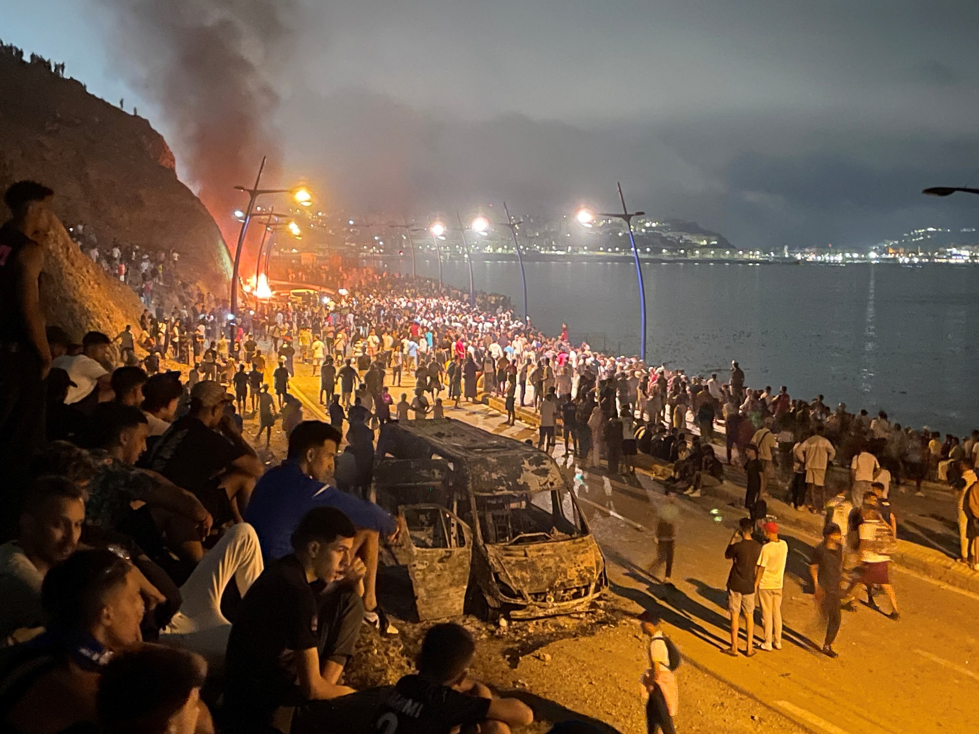A large crowd in Fnideq surrounds the burned shell of a vehicle near the Ceuta border at night