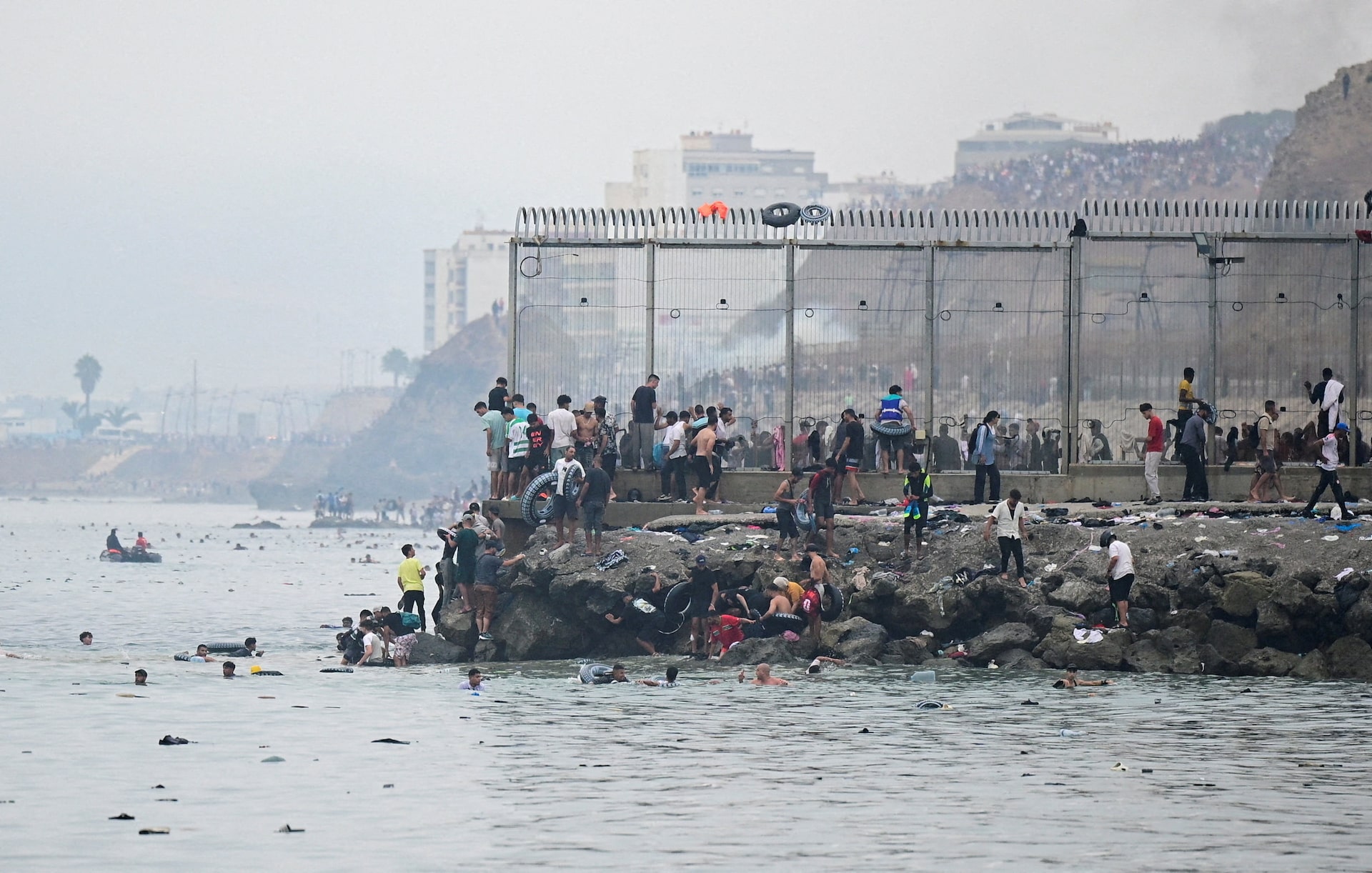 A large crowd gathers at the Ceuta sea border fence and breakwater while swimmers approach through the water